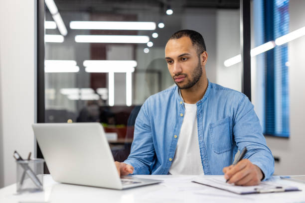 istock Serious young hispanic man working in office at desk with laptop and making notes in notebook, watching online training and meeting 2158446860