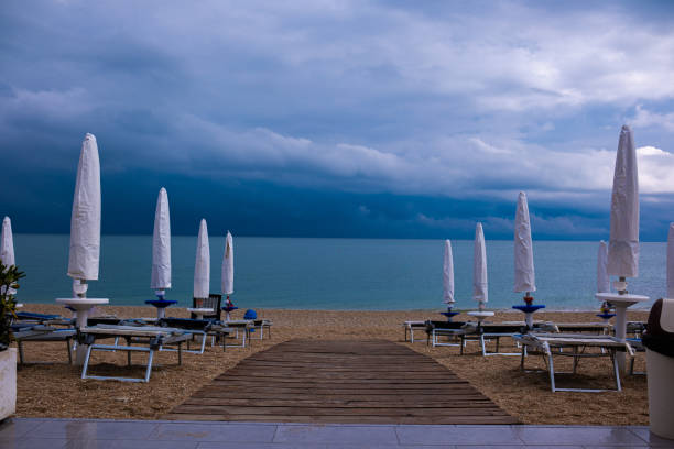 Sea beach in low season. Folded beach umbrellas, deserted paths, empty sunbeds and heavy thunderclouds in the sky above the sea.