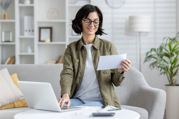 istock Smiling young woman sitting on sofa at home and working on notebook with documents, using laptop and holding papers and bills in hands 2158456517