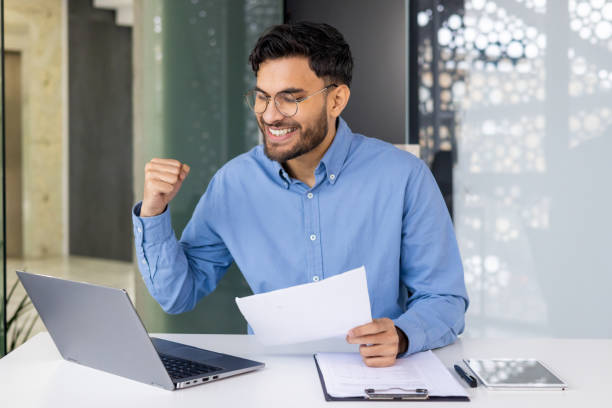 istock Happy businessman celebrating success in office while holding documents and working on laptop 2158459923