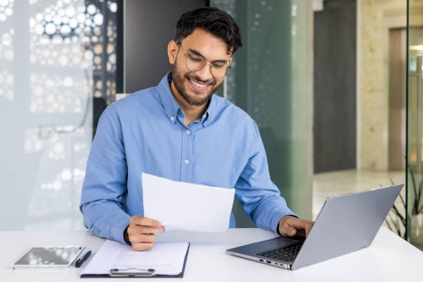istock A smiling young Indian man is sitting in the office at a table with a laptop and working with documents, holding papers and typing data on a keyboard 2158460355