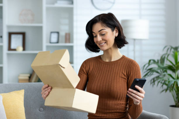 istock A smiling young woman is sitting on the sofa at home, holding a mobile phone and an open cardboard box in her hand, she is happy about the received package and the purchase 2158460607