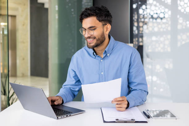 istock Smiling businessman working on laptop and holding paper while sitting in modern office 2158460866