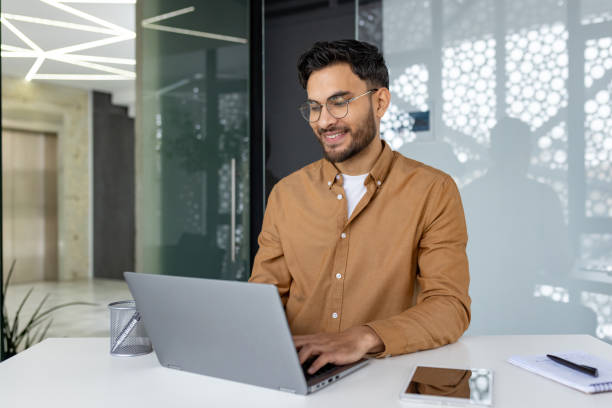 istock Indian young man studying and working in a modern office, sitting at a desk, smiling and typing on a keyboard with concentration 2158461530