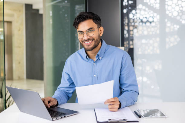 istock Smiling businessman working on laptop and reviewing documents at office desk 2158462586