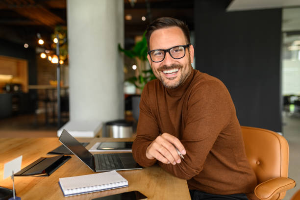 istock Male freelancer with laptop and notepad on desk smiling at camera while sitting on chair in office 2158469714