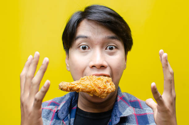 istock A funny, expressive Asian man holds a fried chicken drumstick with a wow expression isolated over a yellow background. 2158483206