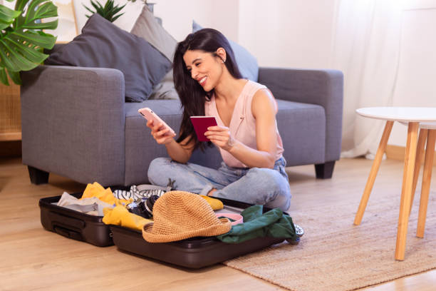 istock Happy woman holding passport using smartphone while packing suitcase on floor at home 2158489400