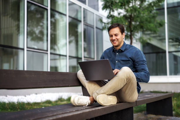 istock Young smiling businessman using laptop while sitting on bench in front of company building 2158492568
