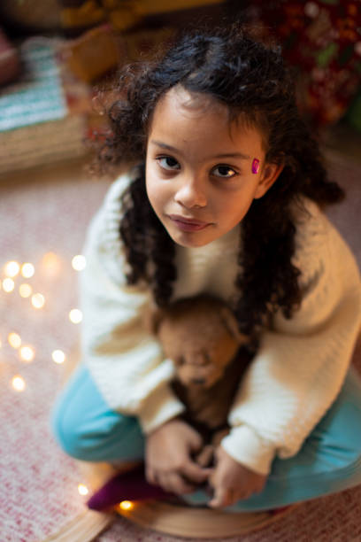 Young girl with a band-aid on her face, holding a teddy bear, sitting indoors during a cozy winter evening, with soft, warm lights in the background.
