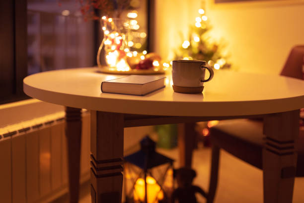 Warm and cozy winter evening inside a home with a book and a mug of tea on a table, soft indoor lighting, and a softly lit Christmas tree in the background, creating a peaceful and relaxing atmosphere.