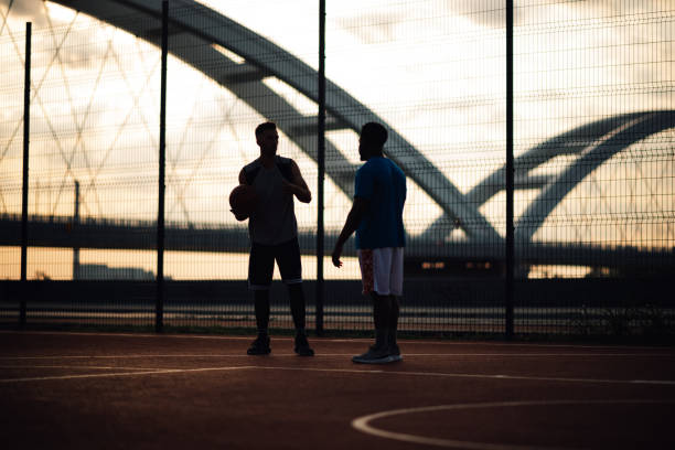 Silhouettes of two men engaged in conversation on a dimly lit basketball court against an evening sky