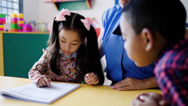 istock Teacher helping kid students learn to read in classroom at kindergarten school room. Multiracial and education concept. Focus on Asian toddler face 2158503354
