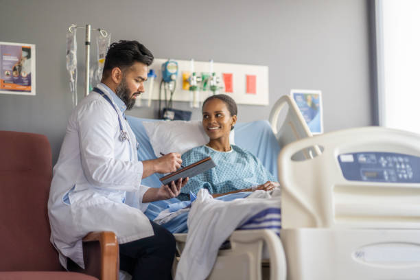 Preparing for Surgery A woman sits up in a hospital bed wearing a gown as she talks with her male doctor about her surgery. The doctor is taking notes electronically on a tablet as the two talk about the procedure. doctor and hospital stock pictures, royalty-free photos & images