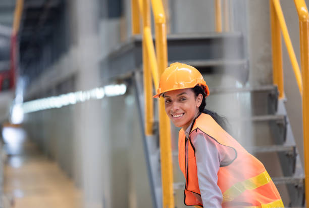 Female engineer with orange hard hat and safety vest is seen from behind, observing train inside maintenance depot. Highlights industrial work, safety, role of women in engineering and transportation