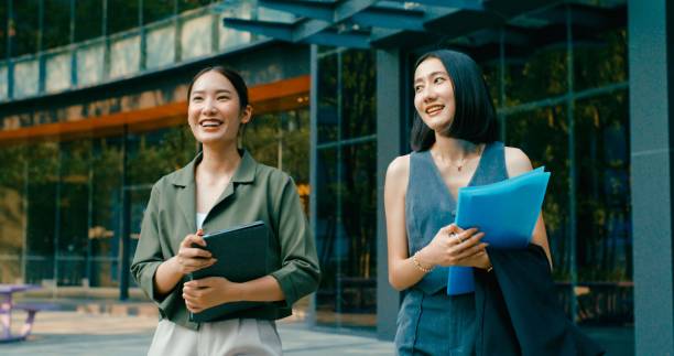 istock Two young Asian businesswomen discussing work while holds digital tablet and carries documents while walking outside office building. Sustainable business outdoor. 2158535861