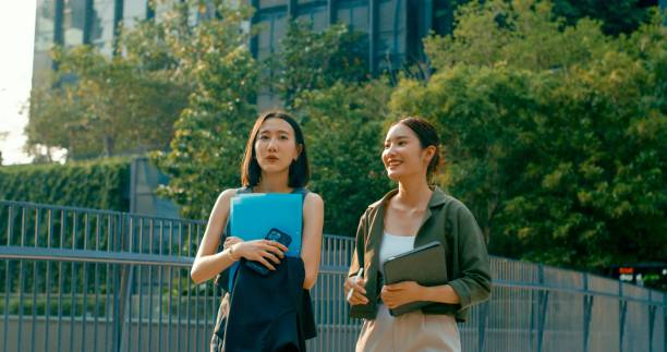 istock Two young Asian businesswomen discussing work while holds digital tablet and carries documents while walking outside office building. Sustainable business outdoor. 2158535923