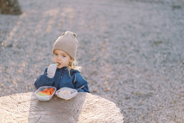 Little girl eats a piece of cucumber while standing behind a large tree stump with a lunchbox. High quality photo