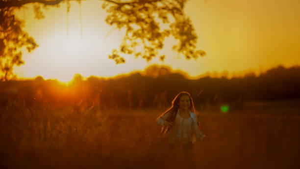A young girl exudes happiness as she runs across a sunlit field, her casual attire fluttering with movement against the warm glow of the evening sky.
