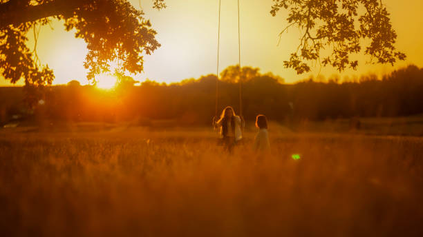 A woman and young girl enjoy a serene swing under a solitary tree as the sun dips low, illuminating the field with a golden hue.