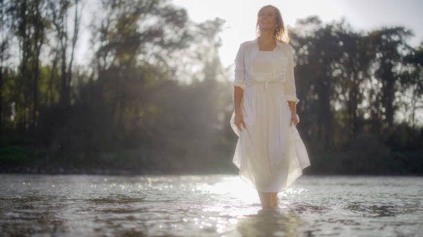 An elegant mature Caucasian woman relaxes in a river, standing against a tranquil backdrop in light, airy attire, embodying peacefulness in a natural setting.