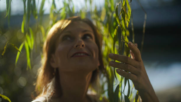A serene woman enjoys the outdoors, gently interacting with the slender leaves of a willow tree, her casual attire blending with the tranquil riverside setting.