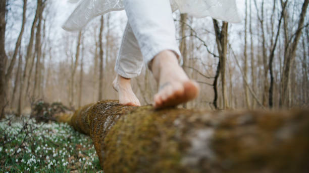 A Caucasian woman expertly treads barefoot on a mossy log amidst a forest blanketed with blooming snowdrops, radiating a serene connection to the natural world around her.