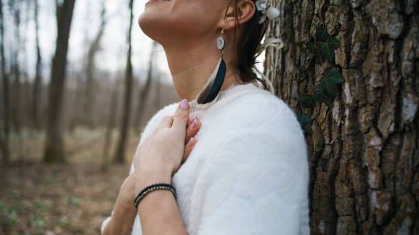 A mature Caucasian woman dressed in a cozy sweater stands by a tree, her posture and gentle clasp of hands signaling gratitude and reflection in the tranquil forest surroundings.