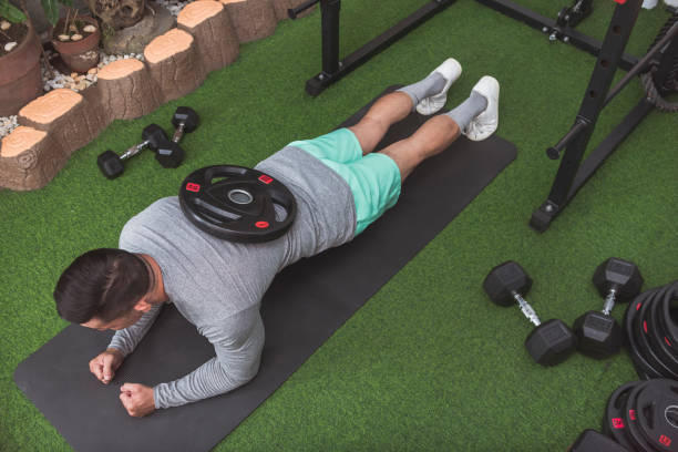 istock A fit asian man doing weighted planks with a barbell plate placed on his back while on a black mat. Core workout at his home gym. 2158547343
