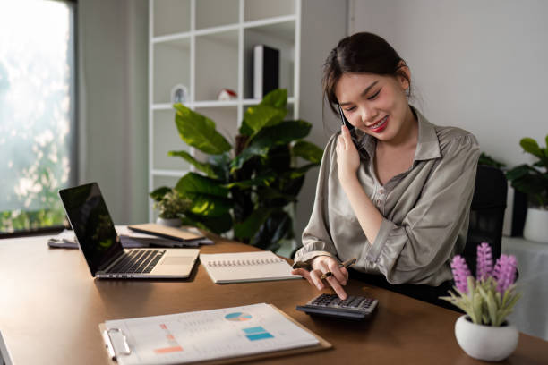 istock Young Business Professional Woman Working from Home Office, Smiling and Using Calculator, Surrounded by Modern Office Supplies, Laptop, and Indoor Plants, in a Bright and Minimalist Workspace 2158564545