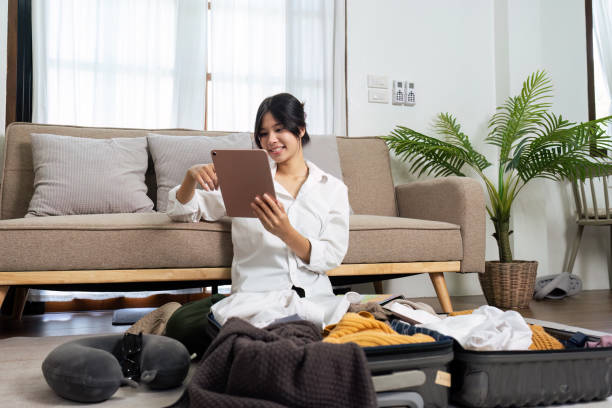 istock Woman Packing Suitcase for Travel While Using Tablet in Modern Living Room with Couch and Plants, Preparing for Vacation or Business Trip, Organizing Clothes and Accessories 2158567013