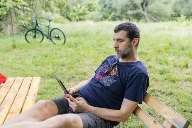 istock Relaxed mid adult man is reading an e-book on digital tablet or e-reader outdoors. He is sitting on wooden bench in caffe in nature. 2158578995
