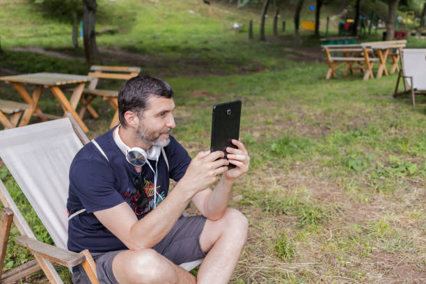 istock Relaxed mid adult man is reading an e-book on digital tablet or e-reader outdoors. He is sitting in comfortable lounge chair in caffe in nature. 2158580200