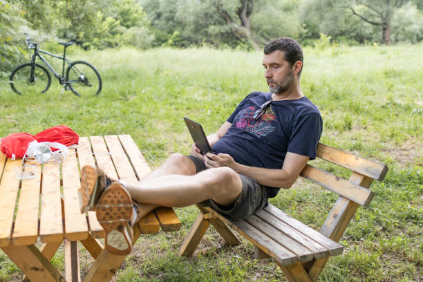 istock Relaxed mid adult man is reading an e-book on digital tablet or e-reader outdoors. He is sitting on wooden bench in caffe in nature. 2158580229