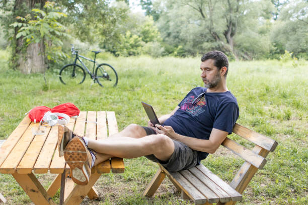 istock Relaxed mid adult man is reading an e-book on digital tablet or e-reader outdoors. He is sitting on wooden bench in caffe in nature. 2158580495