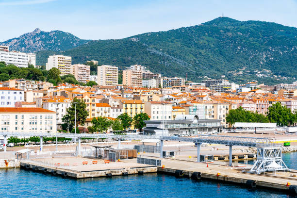 Ajaccio view with a waterfront promenade lined with buildings and a backdrop of hills. The cityscape includes residential and commercial structures, with the marina indicating maritime activities. The hills provide a scenic contrast to the urban setting.