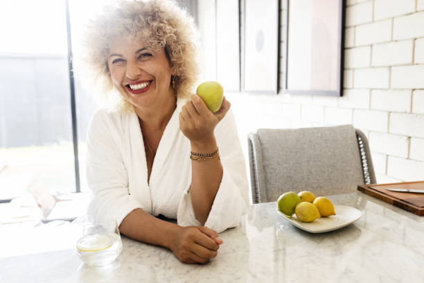 istock Bright and lively image capturing a joyful woman with curly hair, smiling while holding a green apple in a modern, sunlit kitchen setting. A sense of health and positivity emanates from the scene. 2158606489