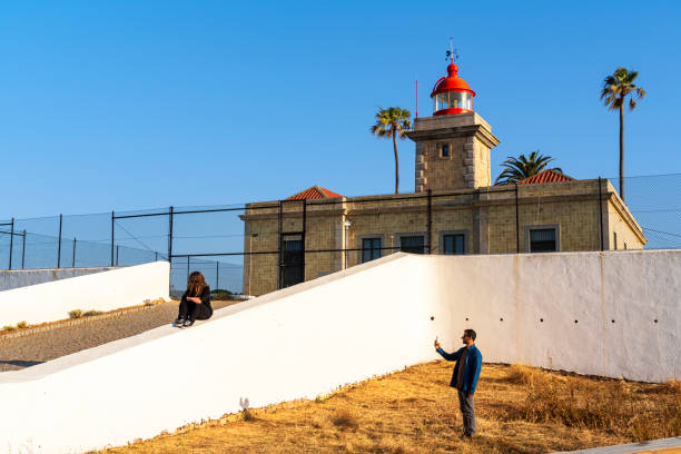 Couple using smartphone to take photos in front of the lighthouse in the morning, Ponta da Piedade, Lagos, Algarve, Portugal