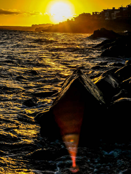 The sun is setting over the ocean, casting a warm glow on the water. The rocks in the foreground create a sense of depth and contrast against the calm sea