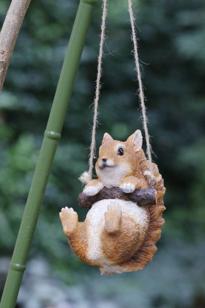 A charming garden ornament of a squirrel hanging from a rustic rope swing. The playful squirrel figure holds onto a small branch with its paws, as if joyfully swinging in mid-air. The background is softly blurred with lush greenery, giving the photo a natural, outdoorsy feel. Perfect for use in themes related to gardening, outdoor decor, or whimsical, playful elements in nature.