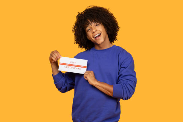 Cheerful Young Man Holding Gift Certificate Against Yellow Background