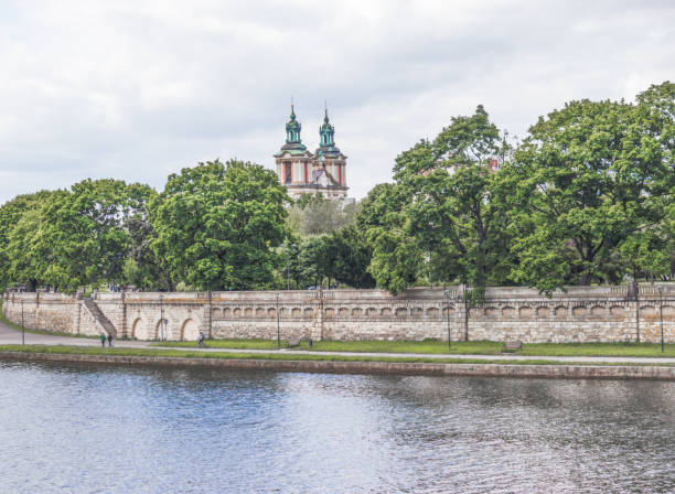 The beautiful river flows along the picturesque bank. The embankment is framed by a mighty ancient stone wall, richly decorated with architectural elements. Behind the wall, huge magnificent trees rise majestically, behind which you can see the ancient church.