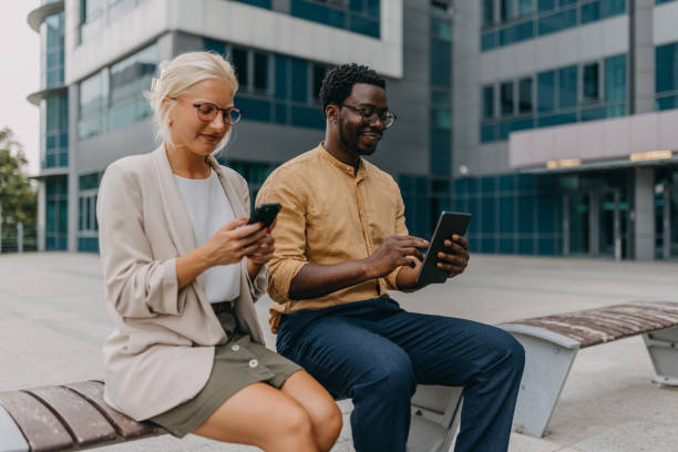 istock Two coworkers on bench using phone and tablet outdoors. 2158655846