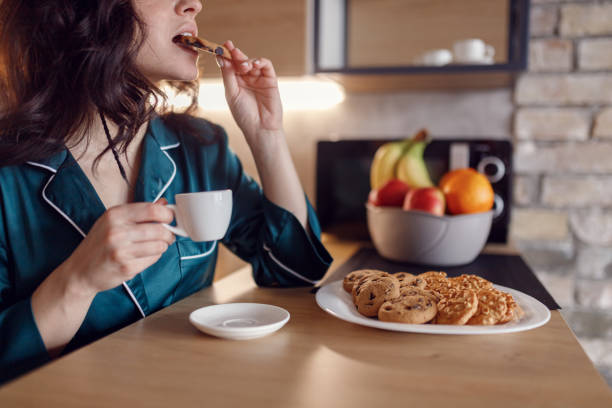 istock Young beautiful woman in silk robe having breakfast at home in the kitchen while holding cup of coffee and biting a cookie. Morning routine, domestic life 2158671106