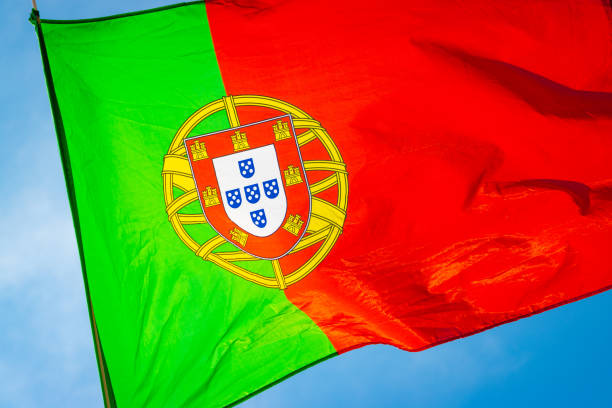 The flag and crest of Portugal backlit by sun flying against a bright blue sky.