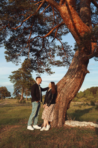 A white guy and a girl are standing by a big pine tree. They are talking and looking at each other.