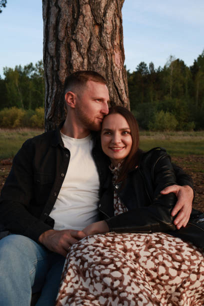A guy and a girl are sitting under a tree. A guy hugs a girl and touches her hand.