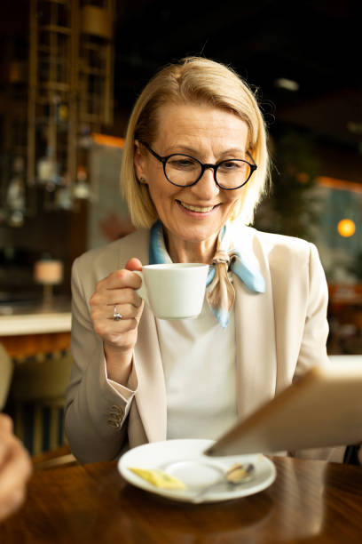 istock Senior woman enjoying coffee while using a tablet in a cozy cafe. She is smiling and appears to be relaxed, indicating the pleasant atmosphere and positive vibes of the place. The scene suggests leisure and comfort. 2158699395