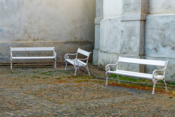Three white benches on a cobblestone street in front of a gray building.