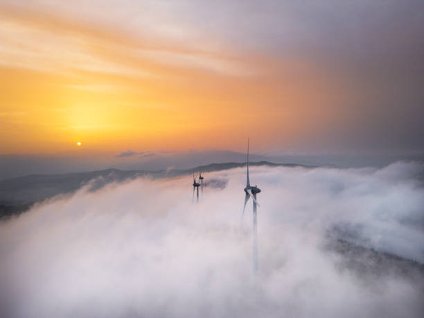 Aerial view of wind turbines emerging through fog at sunrise, with a scenic landscape in the background.